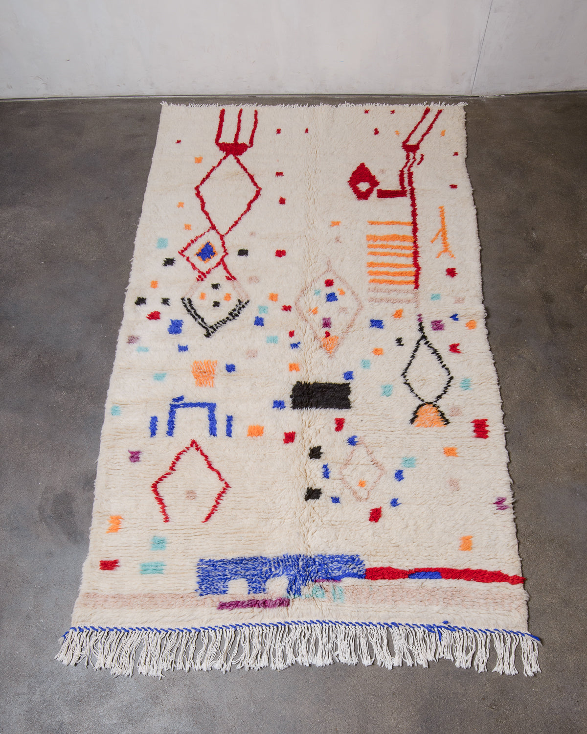 Beniourain Rug made of plush cream wool with a design in red, orange, blue and black laying flat on concrete floor.
